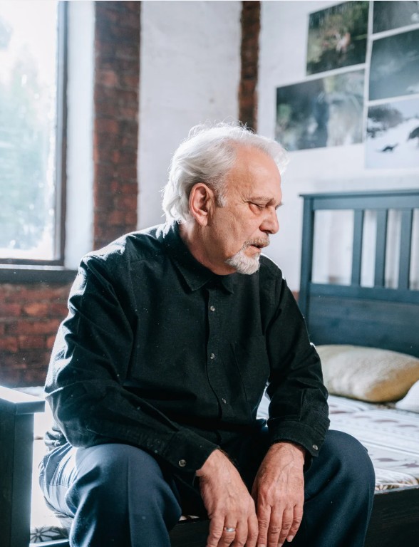 An older man sits on the edge of a bed in a softly lit room, looking down with a reflective, somber expression.