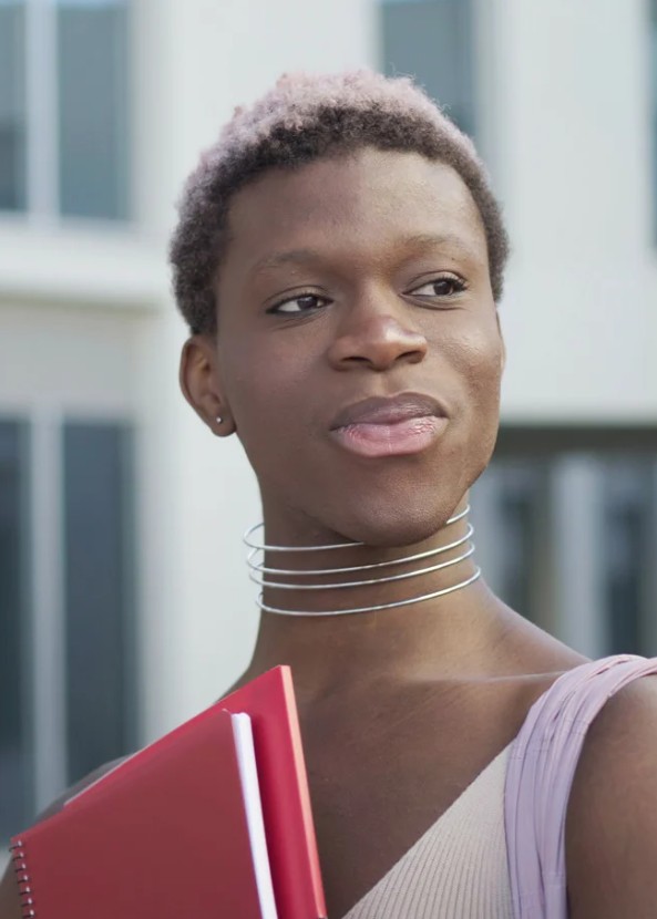 A young adult holding a red notebook looks thoughtfully to the side while standing outdoors near a modern building.